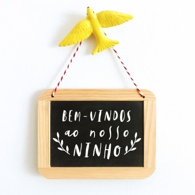 Placa decorativa com moldura de madeira e texto em fundo preto pendurada numa pomba amarela