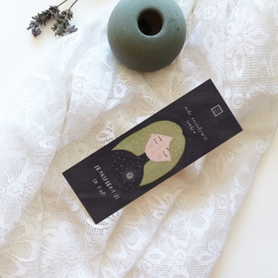 Etiqueta preta com ilustração de mulher e texto sobre tecido branco rendado junto a objeto cerâmico verde.