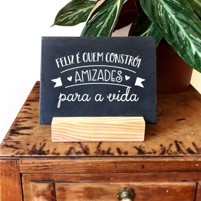 Placa preta com texto branco 'FELIZ É QUEM CONSTRÓI AMIZADES para a vida' em suporte de madeira sobre móvel castanho