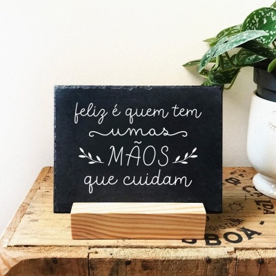 Placa preta com texto branco sobre suporte de madeira ao lado de vaso branco com planta verde