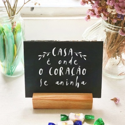 Placa preta com texto branco apoiada em suporte de madeira, entre frascos de vidro com flores e doces coloridos