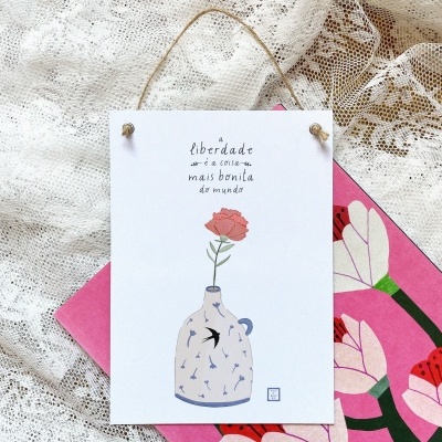 Cartão ilustrado com vaso e rosa pendurado sobre tecido rendado e caderno rosa com flores