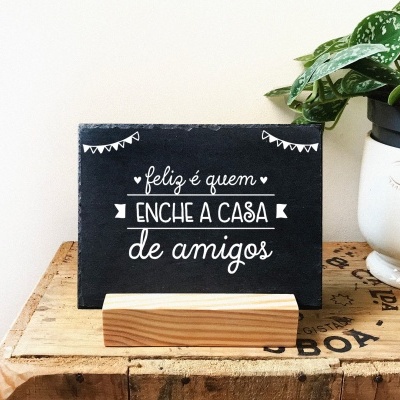 Quadro decorativo preto com texto em branco sobre suporte de madeira ao lado de planta em vaso branco de rosto