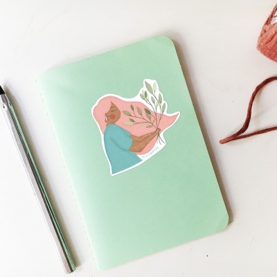 Caderno verde claro com autocolante de mulher a segurar plantas e caneta prateada ao lado.