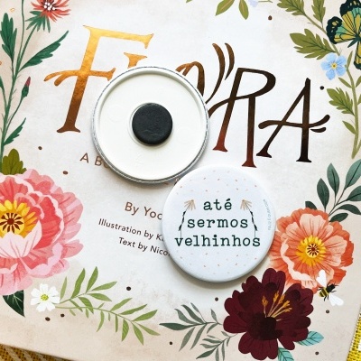 Livro com capa floral e dois bottons brancos, um com texto em português, sobre toalha amarela quadriculada.