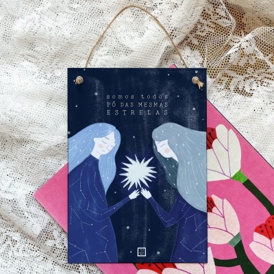Placa decorativa azul com duas figuras femininas segurando uma estrela, com texto em português, sobre fundo de renda branca e bolsa rosa com flores