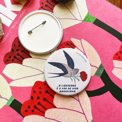 Duas chapas redondas com alfinetes, uma com ilustração de andorinha e texto, sobre tecido florido e revista