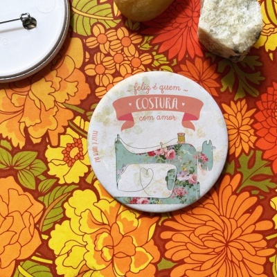 Botão redondo com ilustração de máquina de costura e texto em português sobre tecido floral colorido