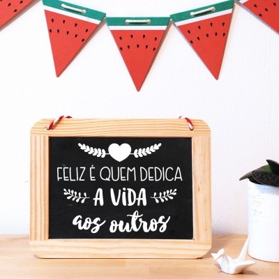 Quadro negro com texto decorativo, vaso com planta e guirlanda de melancia numa mesa de madeira