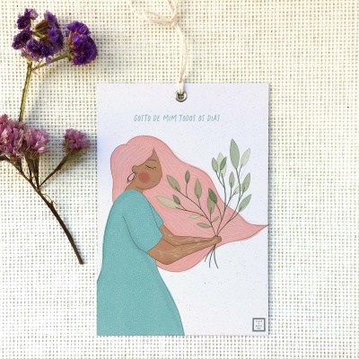 Cartão ilustrado de mulher com vestido azul e cabelo rosa segurando folhas verdes, colocado sobre tecido cru com flores secas ao lado