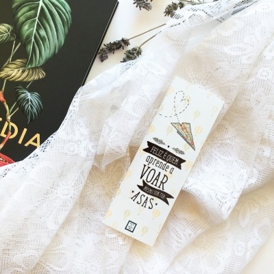 Embalagem branca com texto 'Obrigado por Optares por Voar Asas' sobre tecido rendado, ao lado de livro preto com folhas verdes e texto 'TOPOEDIA'.