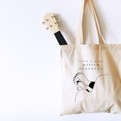 Saco de pano bege com texto e ilustração, contendo ukulele de madeira