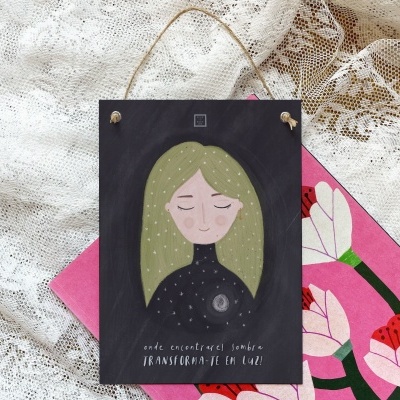 Placa decorativa de madeira com ilustração e texto, pendurada sobre renda e livro.