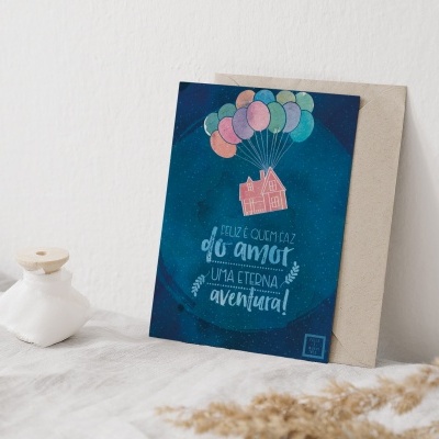 Cartão postal com casa e balões em fundo azul sobre tecido branco, frasco pequeno e ramos de plantas secas ao lado.