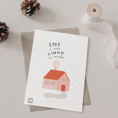 Cartão postal com desenho de casa cor-de-rosa, texto em português, pinhas, envelope castanho e fita branca sobre fundo cinzento claro