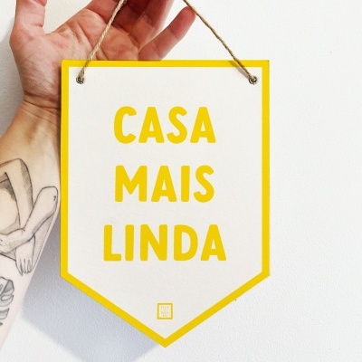 Placa decorativa amarela com texto 'CASA MAIS LINDA' suspensa por fio