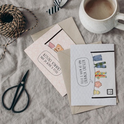 Duas cartas de felicitação, envelopes, tesoura, cordas e caneca sobre tecido bege.
