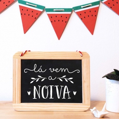 Quadro pequeno em madeira com texto manuscrito, vaso com planta e decoração de bandeirolas em forma de melancia.