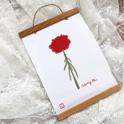 Quadro decorativo com ilustração de flor vermelha simples e moldura de madeira clara sobre tecido de renda branca.