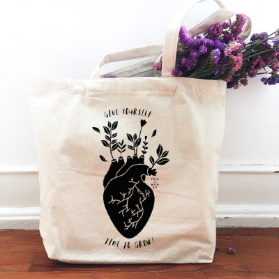 Saco de pano bege com coração anatómico preto e texto, flores lilases dentro.