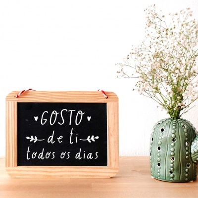 Quadro de madeira com mensagem em português e vaso verde em forma de cacto com flores sobre mesa de madeira