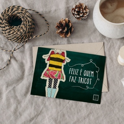Cartão com inscrição FELIZ É QUEM FAZ TRICOT, figura colorida, caneca branca com café e materiais de tricô sobre tecido cinza.