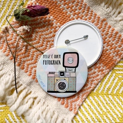 Botom redondo com câmara fotográfica e texto FELIZ É QUEM FOTOGRAFA sobre tecido xadrez com flor seca