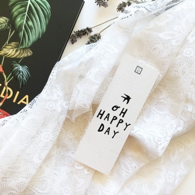 Etiqueta branca com texto 'OH HAPPY DAY' sobre tecido rendado branco, ao lado de livro preto 'TOPEDIA'.