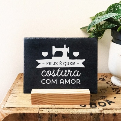 Placa preta com texto branco sobre base de madeira e vaso branco com planta