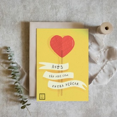 Cartão amarelo com coração vermelho e texto em fita branca sobre superfície cinza