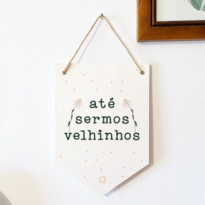 Placa decorativa branca com texto 'até sermos velhinhos' pendurada na parede