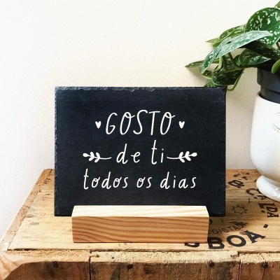 Placa decorativa preta com texto em branco em suporte de madeira, ao lado de vaso branco com planta sobre mesa de madeira