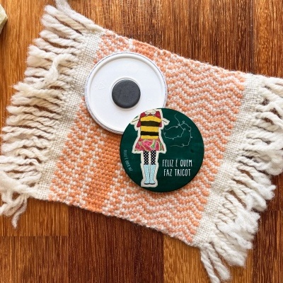 Tapete pequeno com franjas, tampa de plástico branca e íman redondo verde com texto e figura