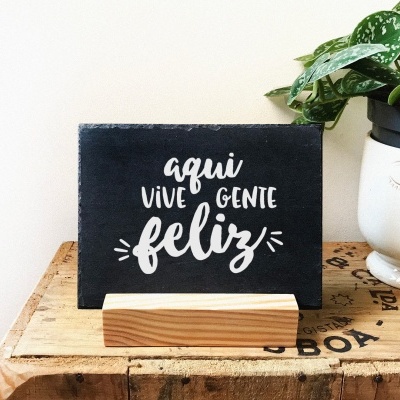 Placa decorativa preta com texto em branco 'aqui vive gente feliz' numa base de madeira ao lado de vaso branco com planta
