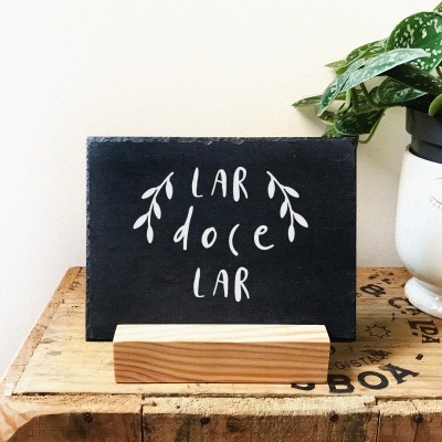 Placa preta escrita 'LAR doce LAR' com suporte de madeira ao lado de vaso branco com plantas
