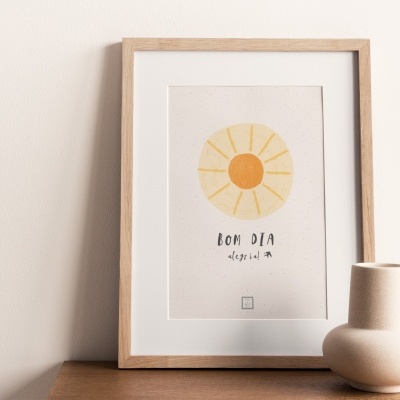 Quadro com ilustração de sol e texto 'BOM DIA' numa moldura de madeira clara junto a vaso branco e esfera de pedra