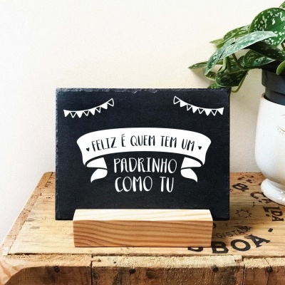 Placa decorativa preta com texto branco 'FELIZ É QUEM TEM UM PADRINHO COMO TU' em suporte de madeira, sobre mesa rústica, junto a planta em vaso branco com rosto.