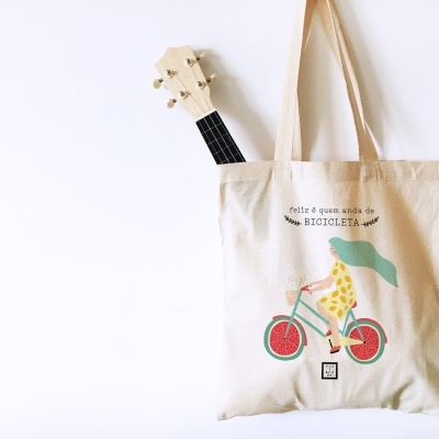 Saco de pano bege com ilustração e texto, pendurado numa parede branca, com um ukulele dentro.