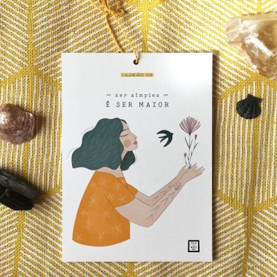 Calendário de papel com ilustração de mulher e texto em fundo amarelo com pedras
