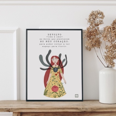 Pôster ilustrado com mulher e texto emoldurado a preto numa mesa de madeira junto a vaso branco com flores secas.