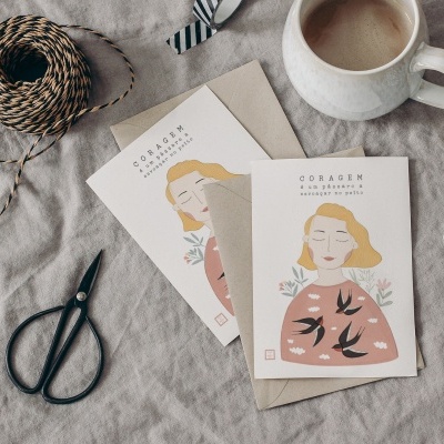 Cartões cor-de-rosa com ilustrações de mulher e texto CORAGEM junto a envelope, tesouras, rolos de fio e caneca em fundo cinzento
