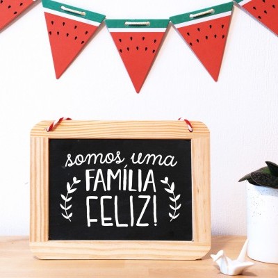 Quadro com texto 'somos uma FAMÍLIA FELIZ!' pendurado numa parede branca, decoração de melancias e vaso com planta suculenta