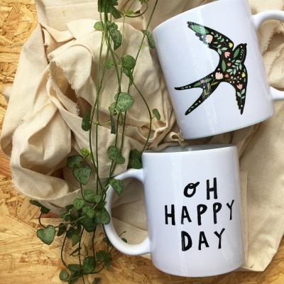 Duas canecas brancas, uma com pássaro colorido e outra com texto 'OH HAPPY DAY' sobre tecido bege e madeira
