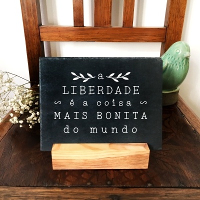Placa decorativa preta com texto branco em suporte de madeira sobre cadeira de madeira escura com flores brancas e figura de pássaro verde