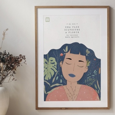 Quadro emoldurado com ilustração de rosto feminino e plantas em fundo azul, pendurado na parede ao lado de um vaso branco com planta seca