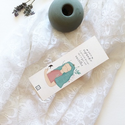 Marca páginas com mulher ilustrada, texto em inglês, sobre pano rendado branco e vaso verde fosco