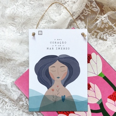 Placa decorativa com ilustração e texto, tecido de renda branca e caderno cor-de-rosa com flores