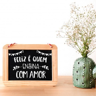 Quadro com a frase FELIZ É QUEM ENSINA COM AMOR e vaso cacto com flores sobre mesa de madeira