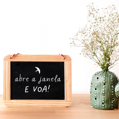 Placa preta com moldura de madeira e vaso em forma de cacto com flores sobre superfície de madeira.