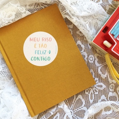 Caderno mostarda com texto em etiqueta branca e câmara Fisher-Price vermelha sobre tecido rendado branco